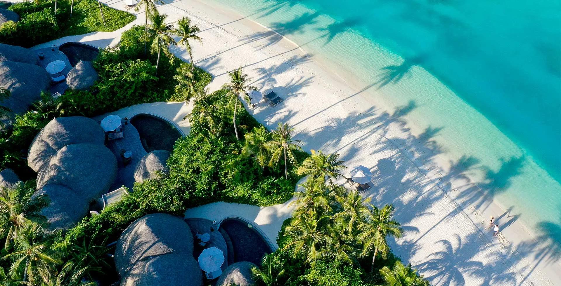 Aerial view of Nautilus Maldives' thatched-roof houses along the beach, palm shadows reaching toward the turquoise lagoon.