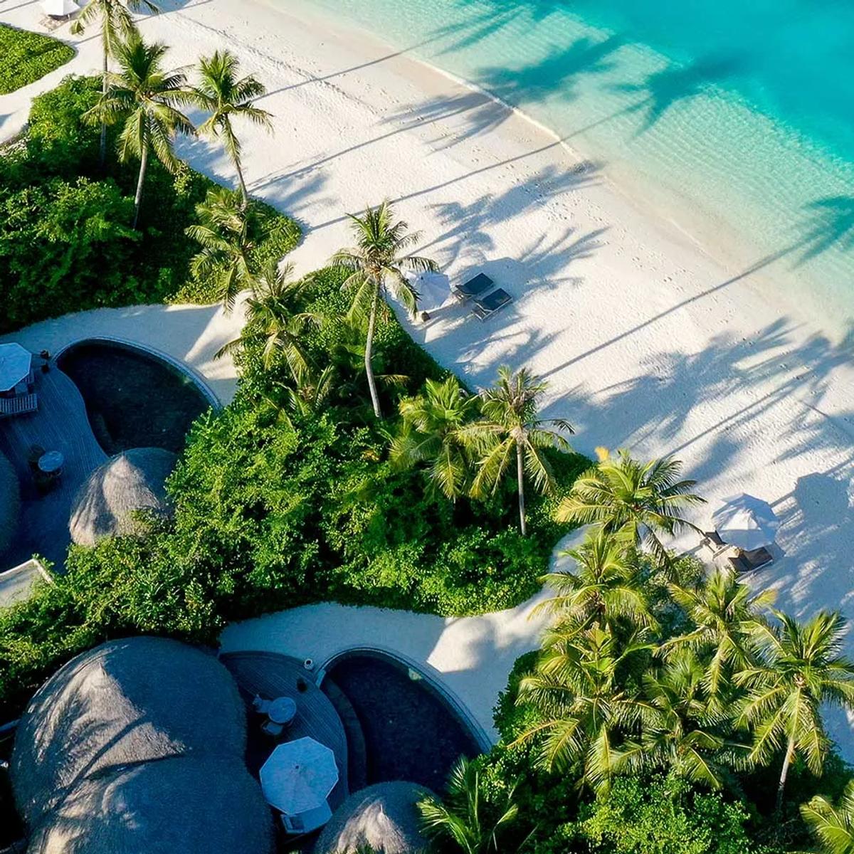Aerial view of Nautilus Maldives' thatched-roof houses along the beach, palm shadows reaching toward the turquoise lagoon.