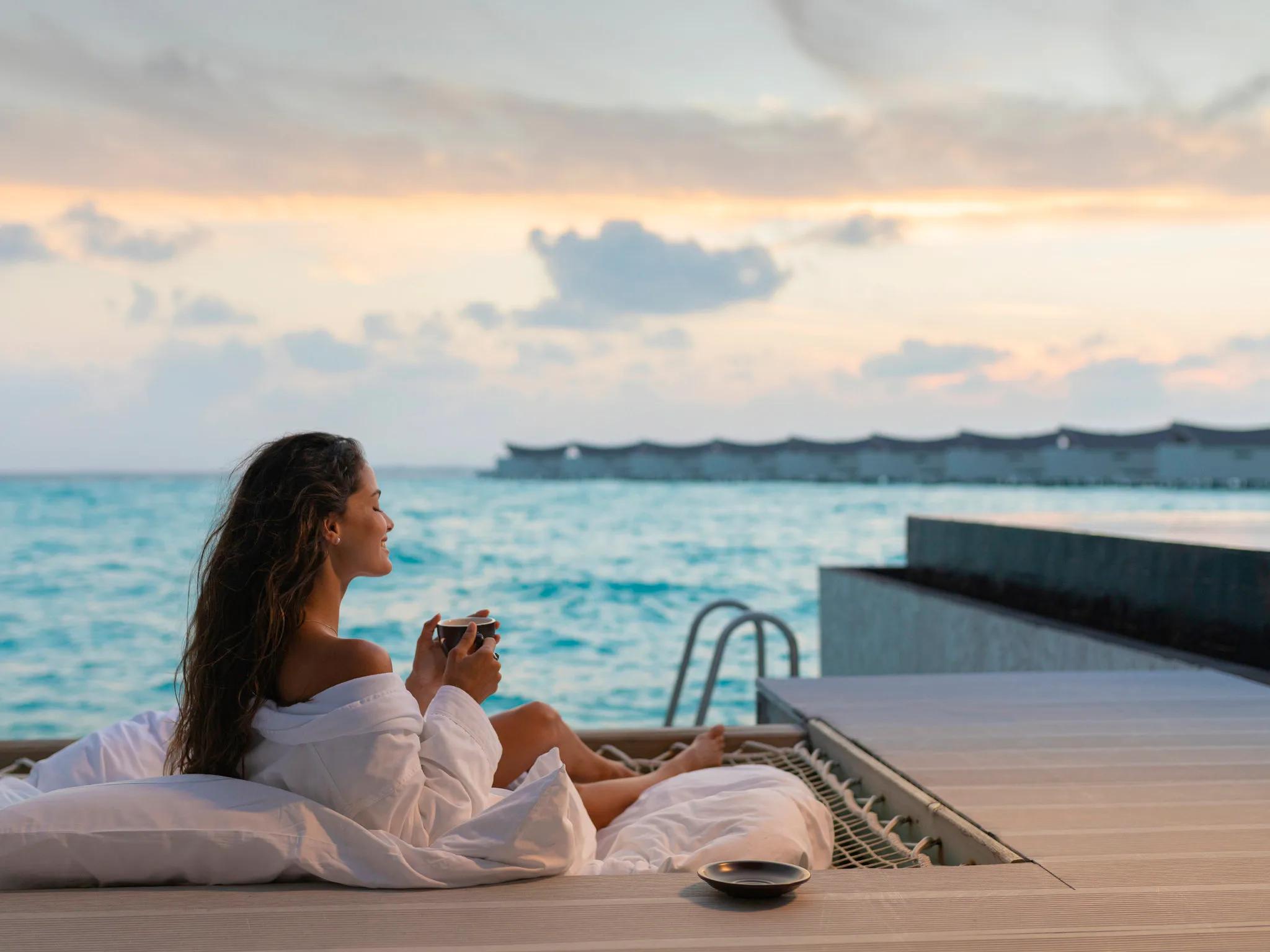 A woman in a white robe drinking coffee on an overwater deck at dusk, a row of pointed-roof villas curving across the lagoon behind her