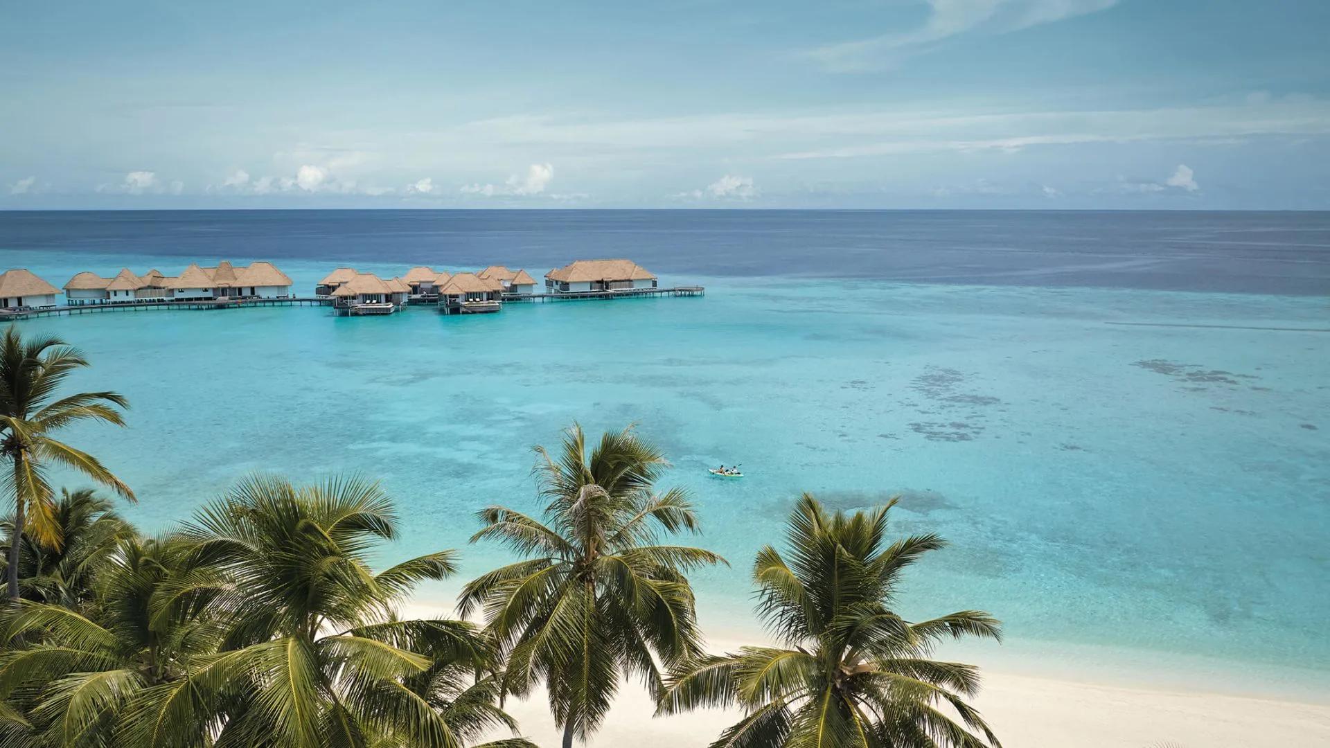 Elevated view through palm trees on the beach, looking out to a row of overwater villas across the lagoon, with two guests kayaking in the shallows