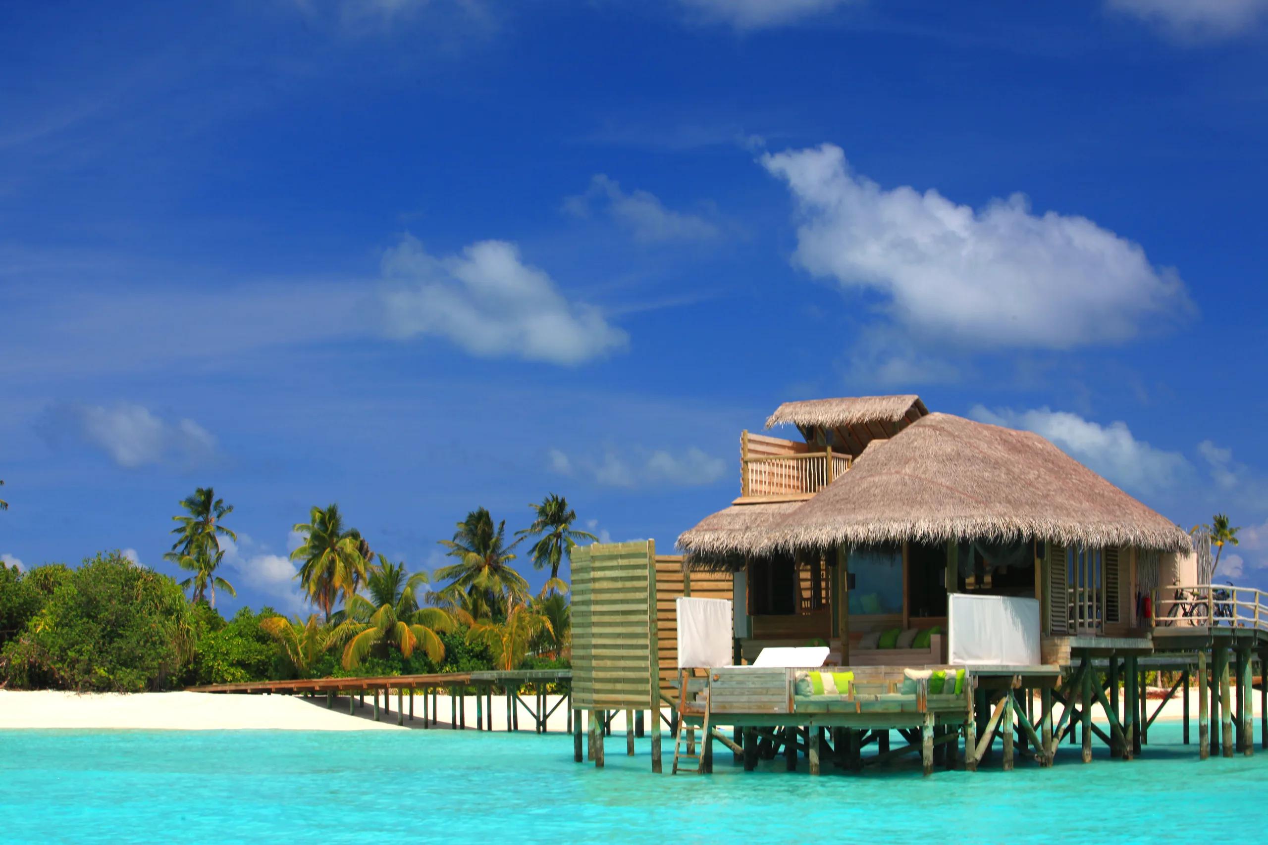 A thatched-roof water villa on stilts above turquoise lagoon water, palm trees and beach behind it