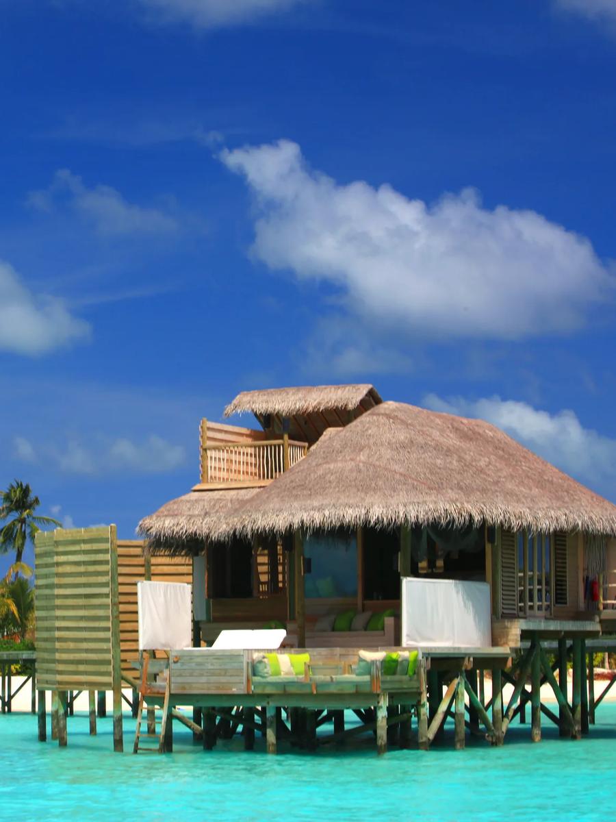 A thatched-roof water villa on stilts above turquoise lagoon water, palm trees and beach behind it