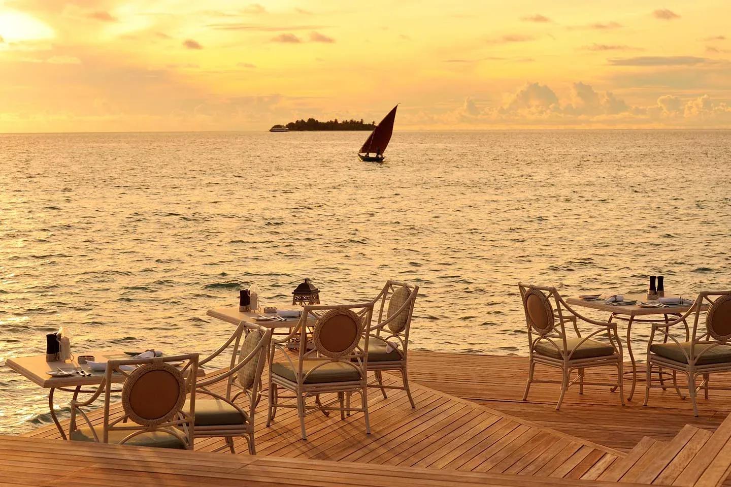 Ocean Breeze's overwater deck at sunset, a red-sailed dhoni passing on the horizon
