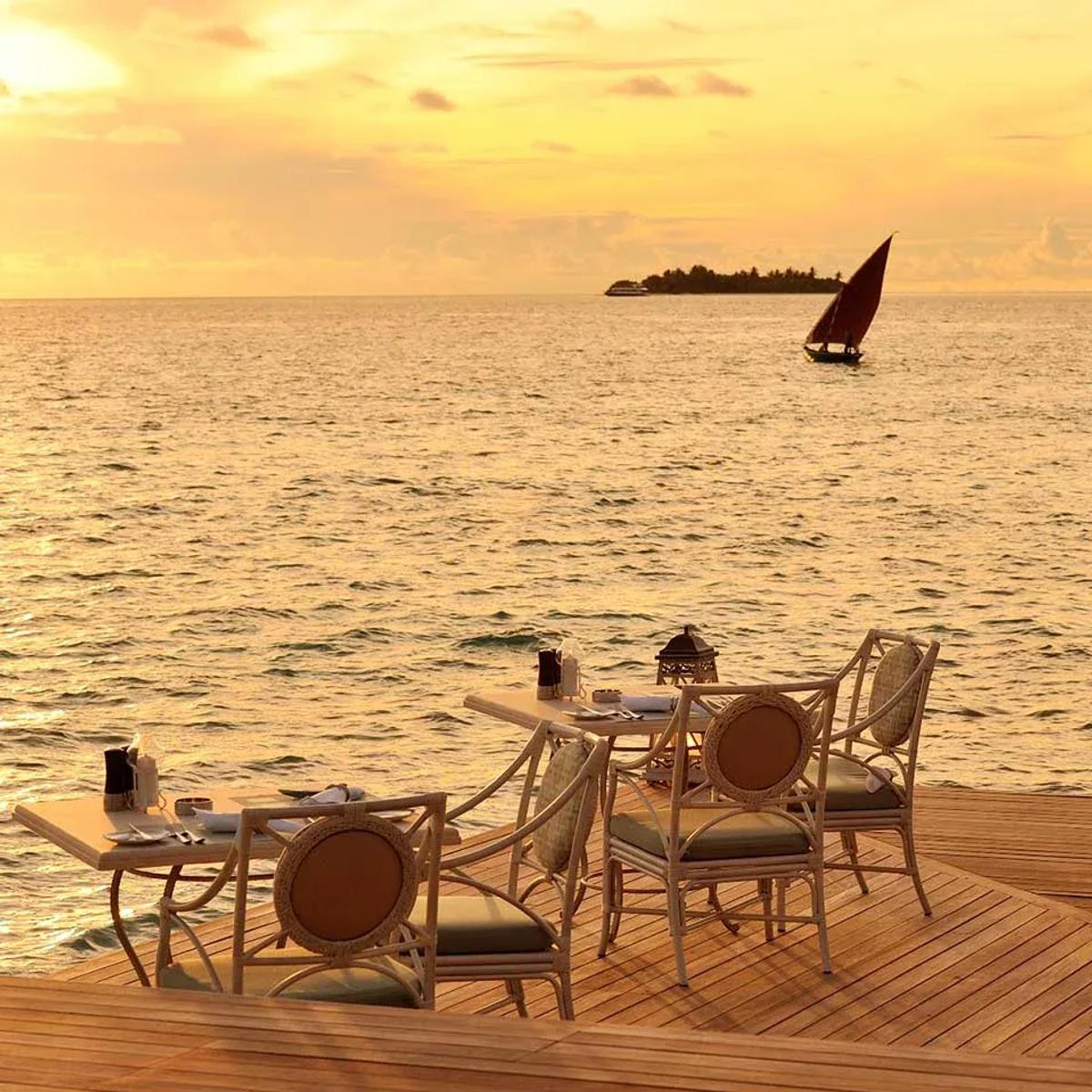 Ocean Breeze's overwater deck at sunset, a red-sailed dhoni passing on the horizon