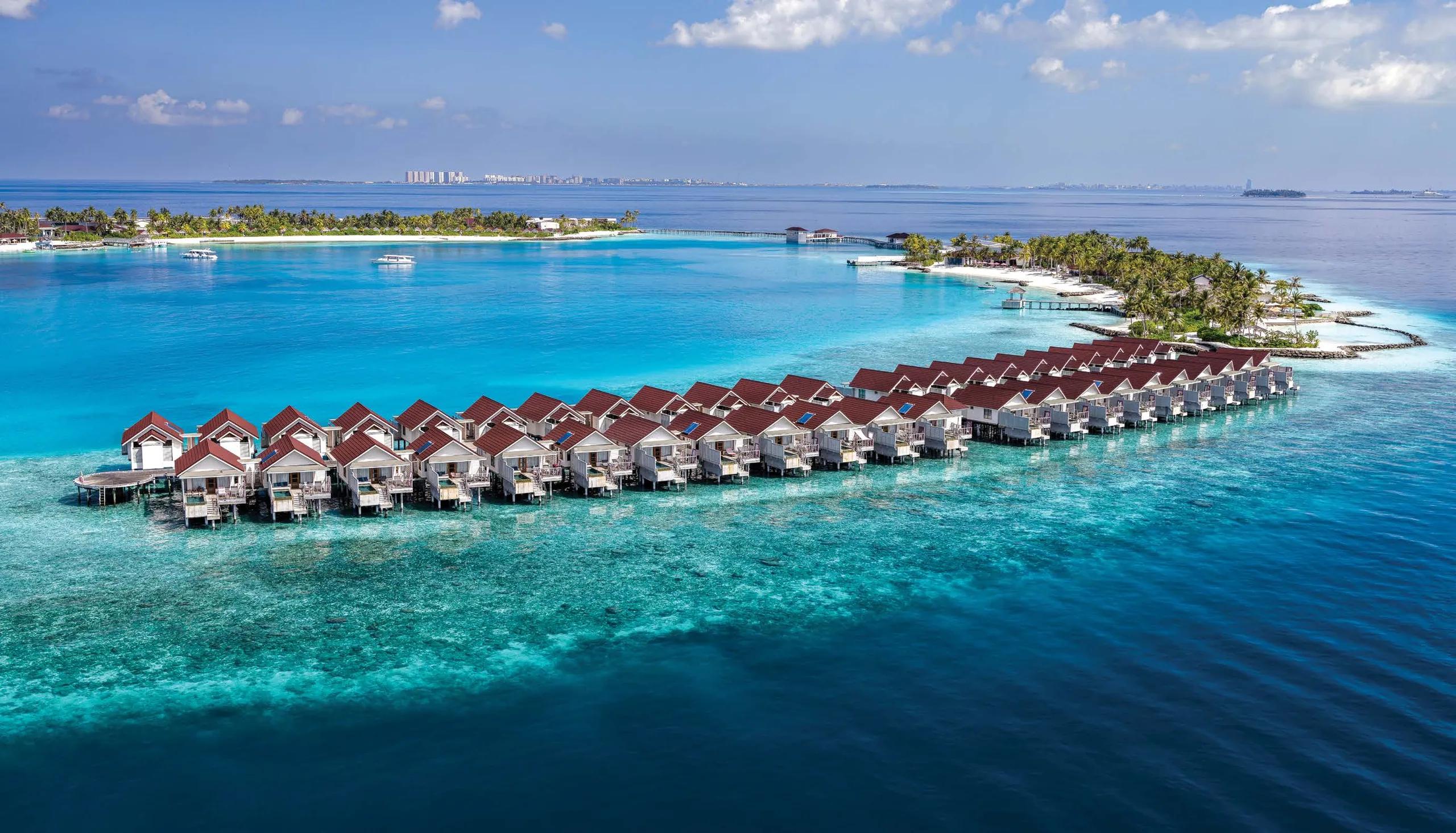 Aerial view of OBLU SELECT Lobigili's overwater villa jetty reaching into the North Malé Atoll lagoon, Malé's skyline visible on the horizon
