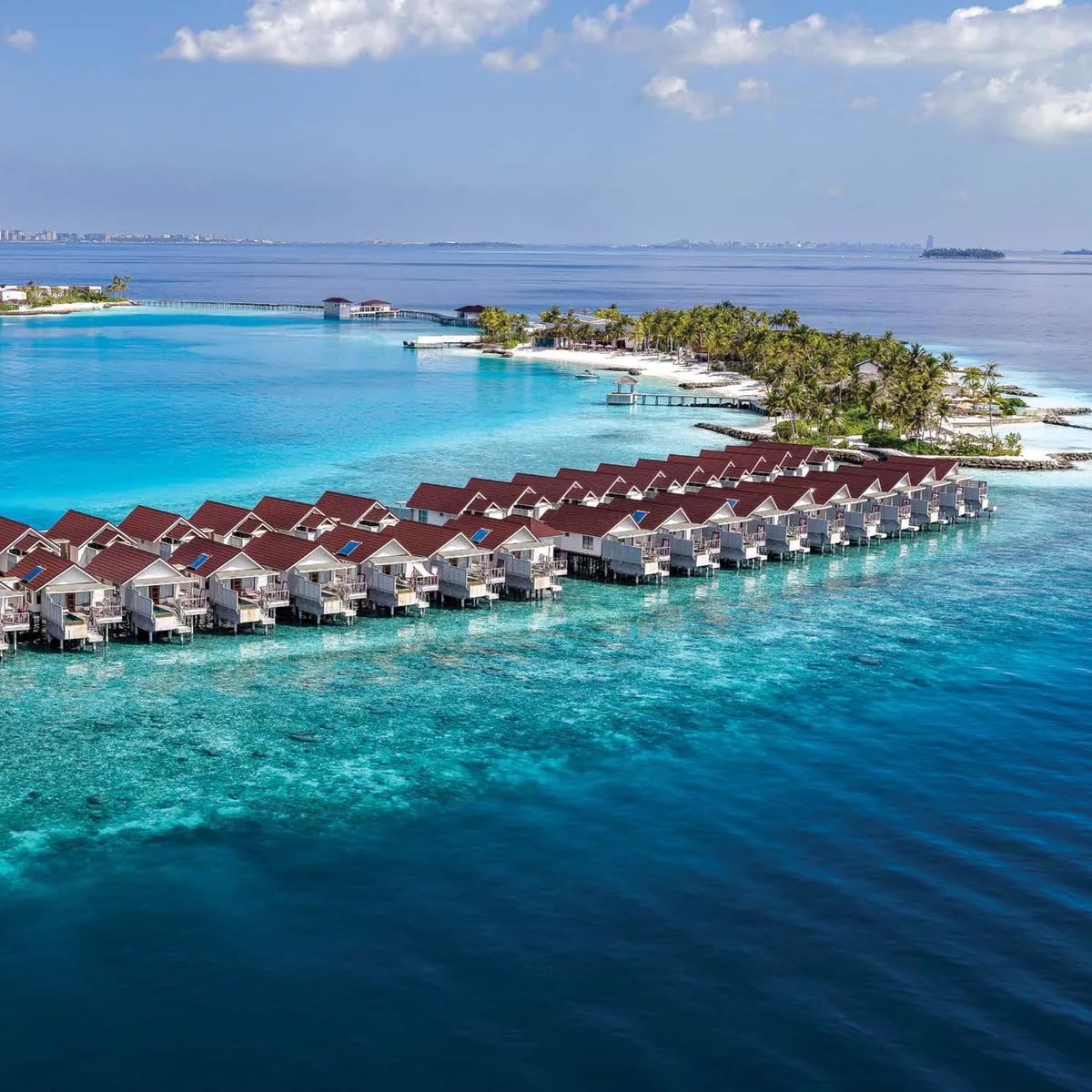 Aerial view of OBLU SELECT Lobigili's overwater villa jetty reaching into the North Malé Atoll lagoon, Malé's skyline visible on the horizon