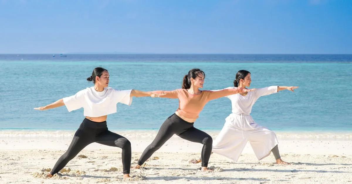 Three women in a beachfront yoga class at Dusit Thani Maldives, arms outstretched in warrior pose facing the sea
