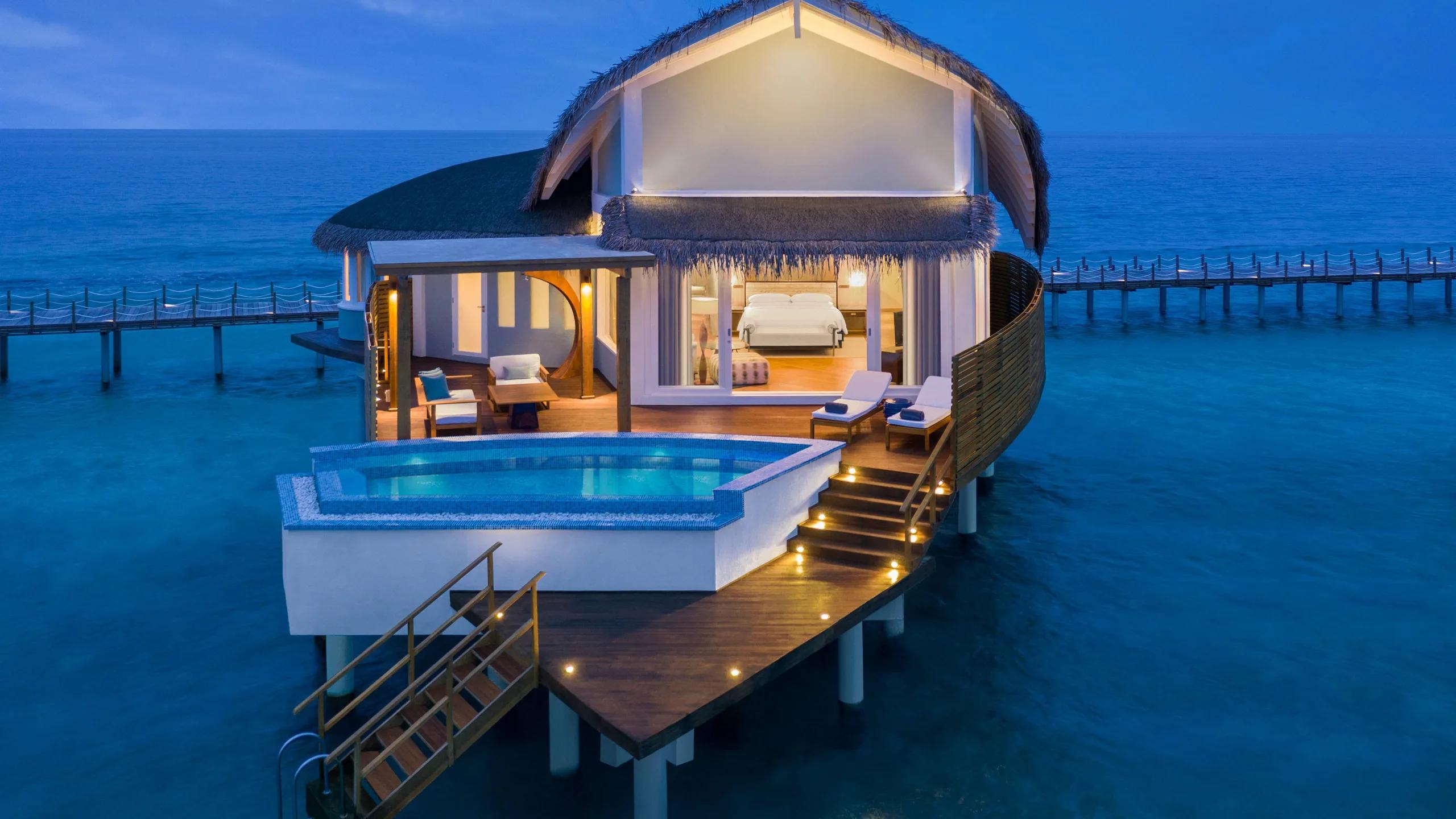 An Overwater Pool Villa at dusk, its private pool and deck lit up above the lagoon, bedroom visible through the glass doors