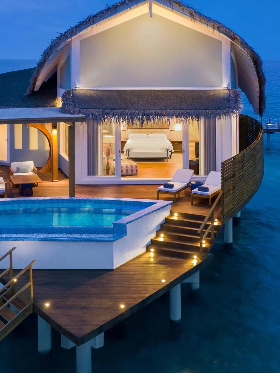 An Overwater Pool Villa at dusk, its private pool and deck lit up above the lagoon, bedroom visible through the glass doors