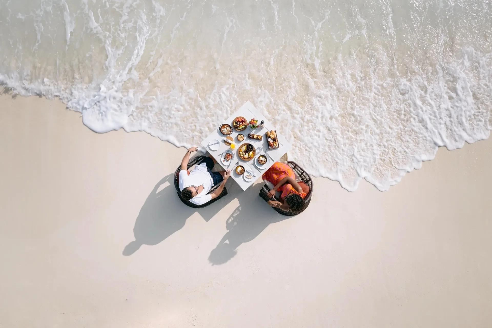 Aerial view of two guests at a table set directly on the sand at the surf line, breakfast plates laid out between them