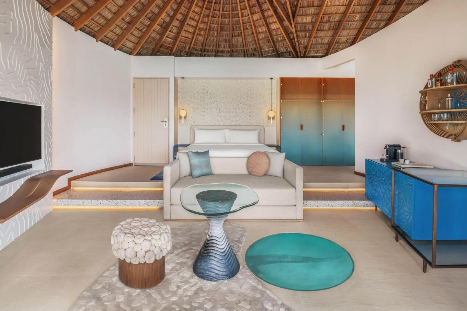 The bedroom of a Superior Overwater villa at W Maldives, under a round thatched roof with a blue-and-orange colour palette
