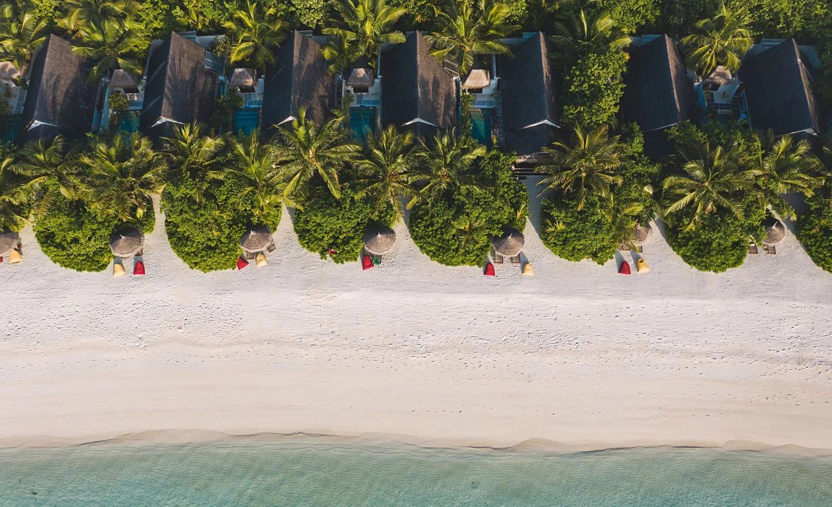 Aerial view of Ozen Life Maadhoo's beachfront villas, thatched roofs set among palms beside the turquoise lagoon