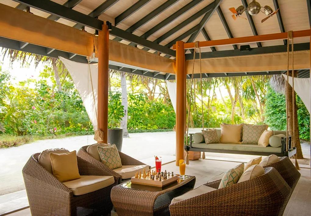 Sails Bar's open-air wooden cabana lounge with woven armchairs, a swinging daybed and a chess set, framed by coconut palms