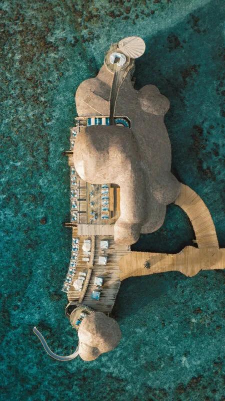 An aerial view of Out of the Blue's overwater dining pavilion, its lobed roofline built out over the reef and connected to the island by a long boardwalk