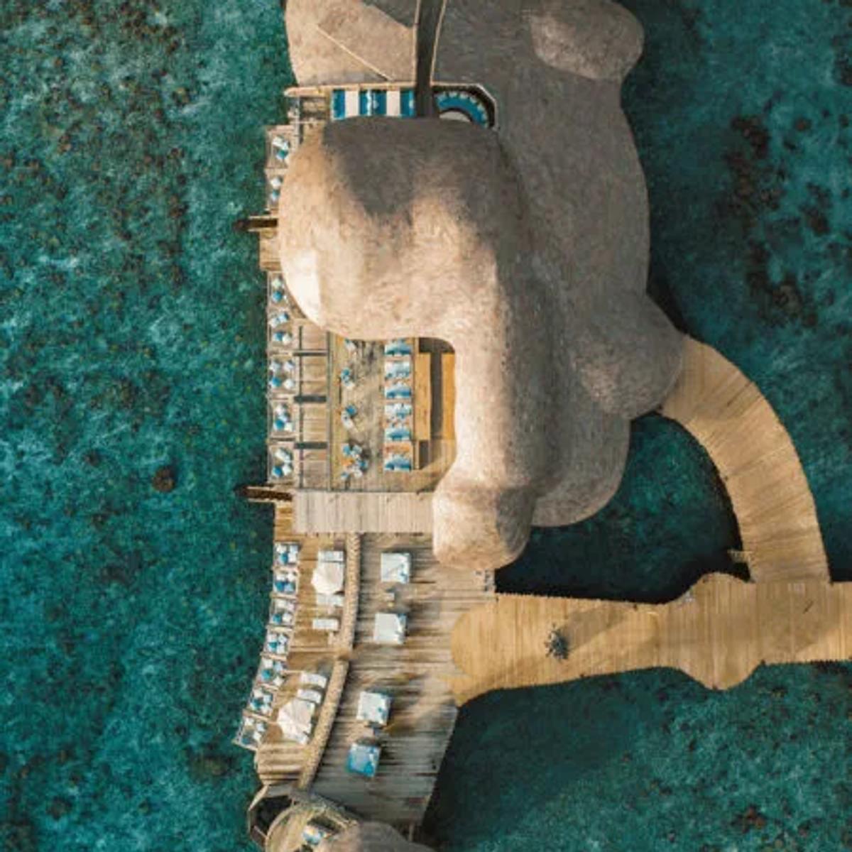 An aerial view of Out of the Blue's overwater dining pavilion, its lobed roofline built out over the reef and connected to the island by a long boardwalk