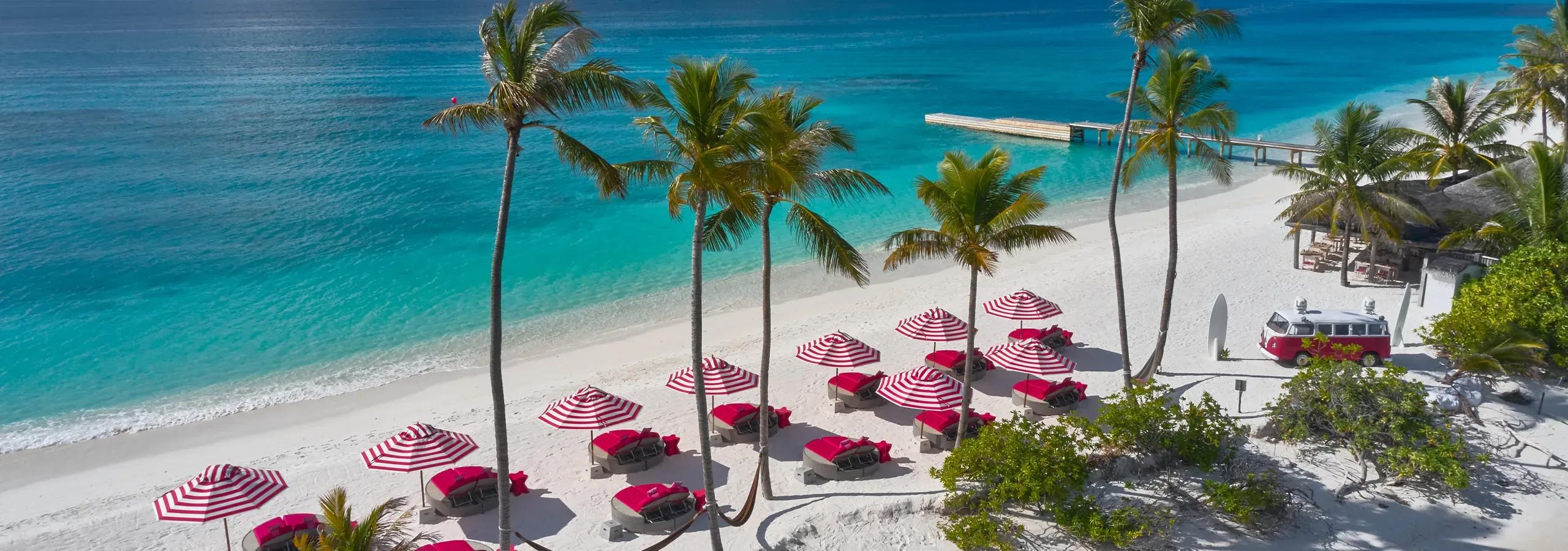 An elevated view of Beach Club's stretch of beach, with red-and-white striped umbrellas over pink loungers, a wooden jetty into the lagoon, and a red vintage VW van parked among the palms