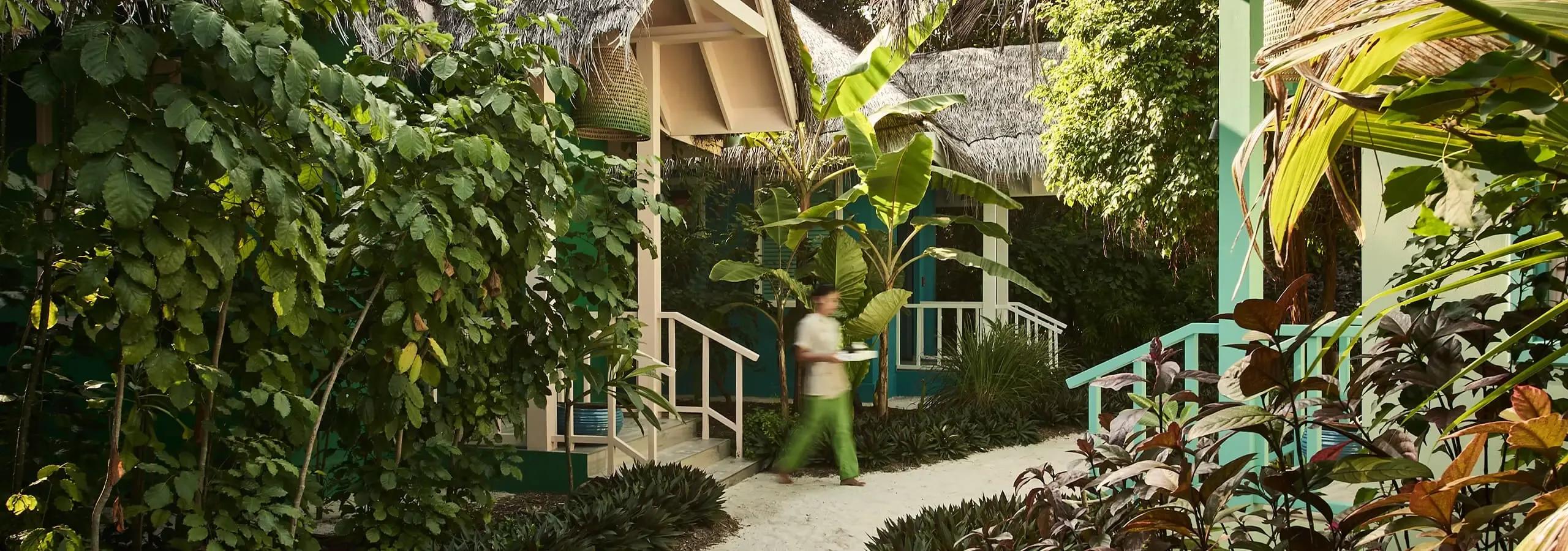 A sandy garden path lined with banana palms and teal-painted buildings, a staff member carrying a tray past Fehi Spa's treatment pavilions