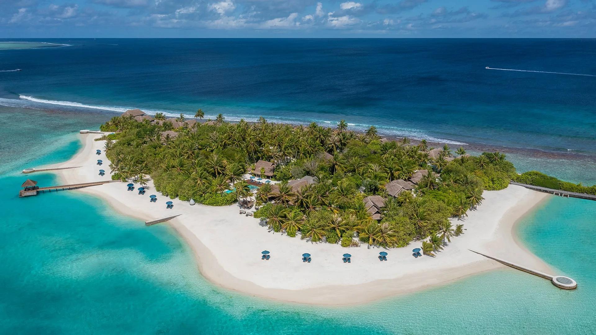 A sweeping aerial view of Naladhu Private Island, its thatched-roof houses set among dense palms on a white-sand island ringed by reef in South Malé Atoll