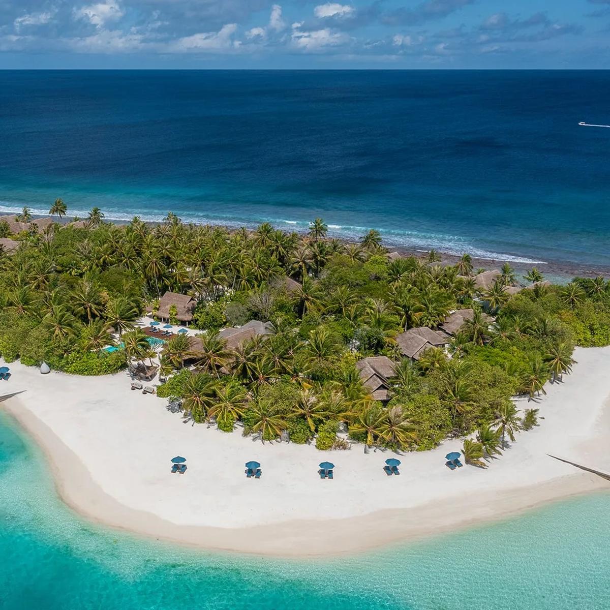 A sweeping aerial view of Naladhu Private Island, its thatched-roof houses set among dense palms on a white-sand island ringed by reef in South Malé Atoll