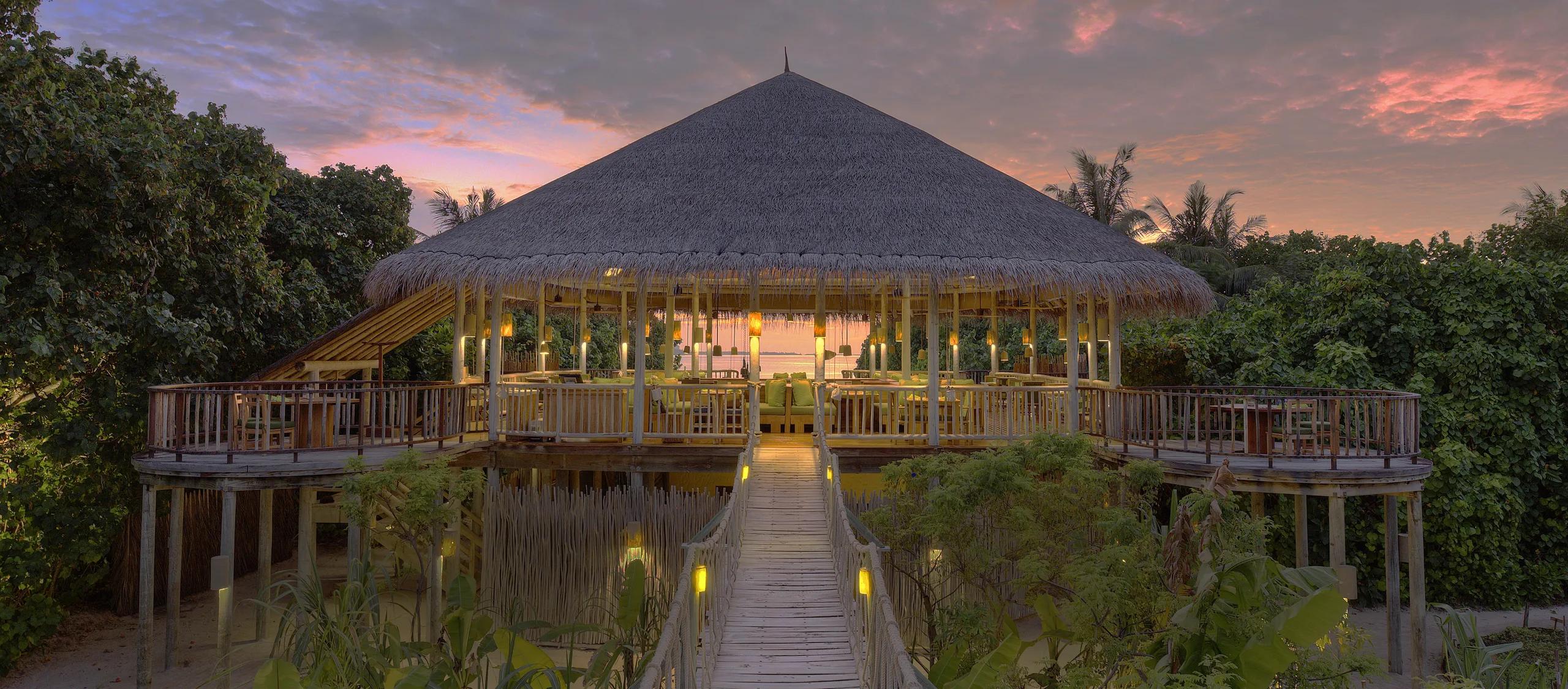 An open-air dining deck under a thatched roof at sunset, with a reef break visible in the distance