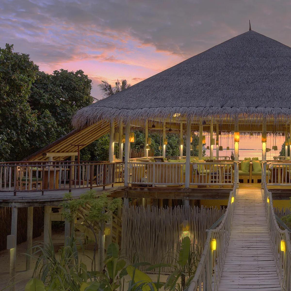 An open-air dining deck under a thatched roof at sunset, with a reef break visible in the distance