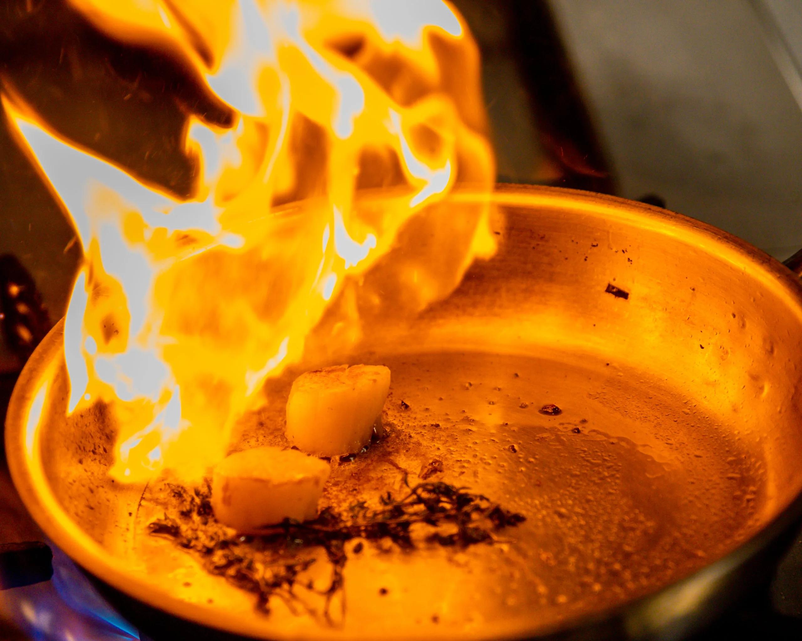 A chef searing scallops tableside, flames rising from the pan over sprigs of thyme