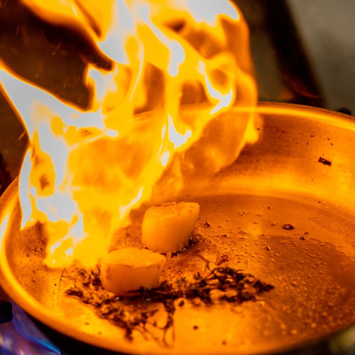 A chef searing scallops tableside, flames rising from the pan over sprigs of thyme