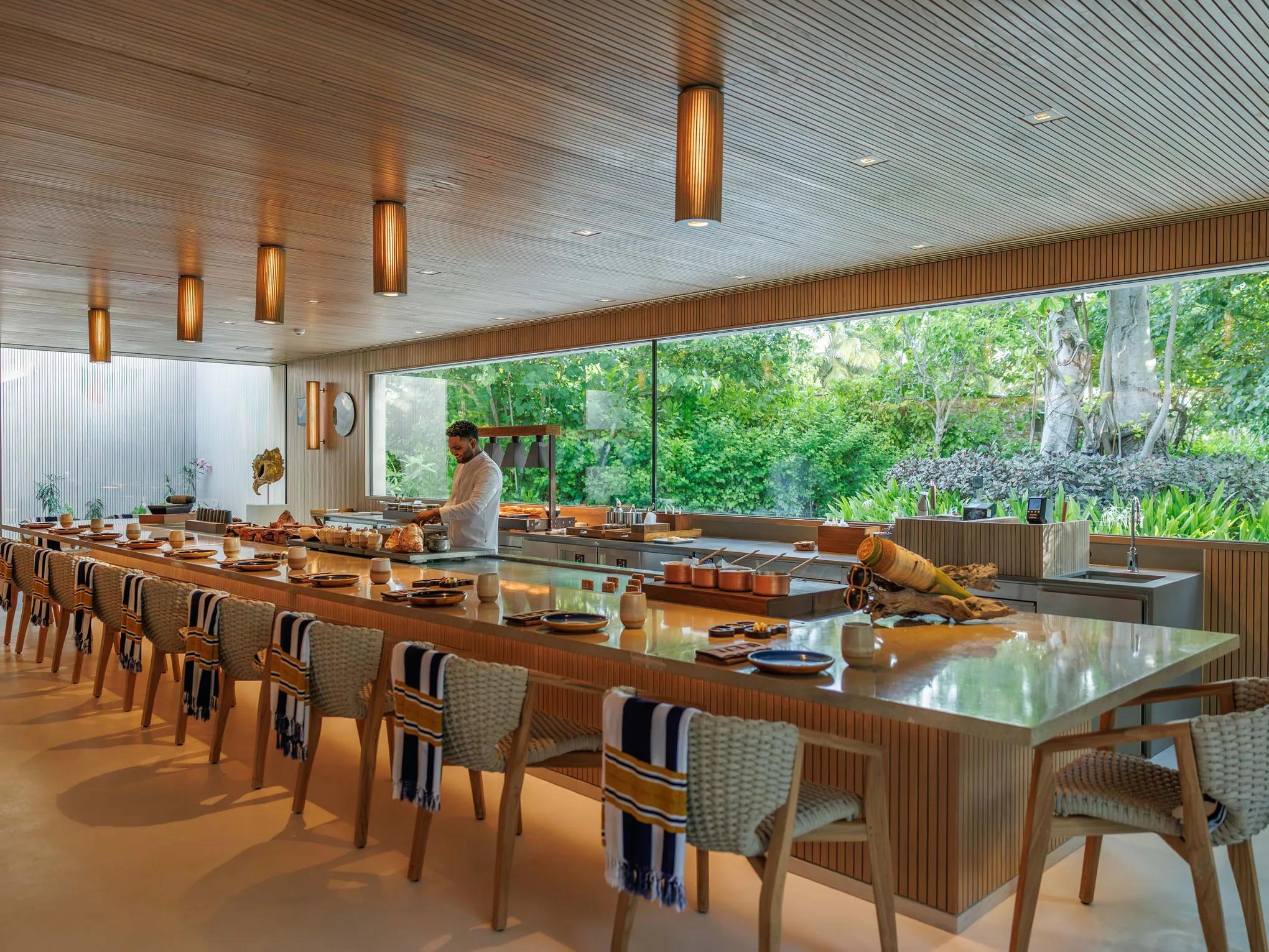 A chef preparing a dish at an open kitchen counter, floor-to-ceiling glass framing the garden behind him