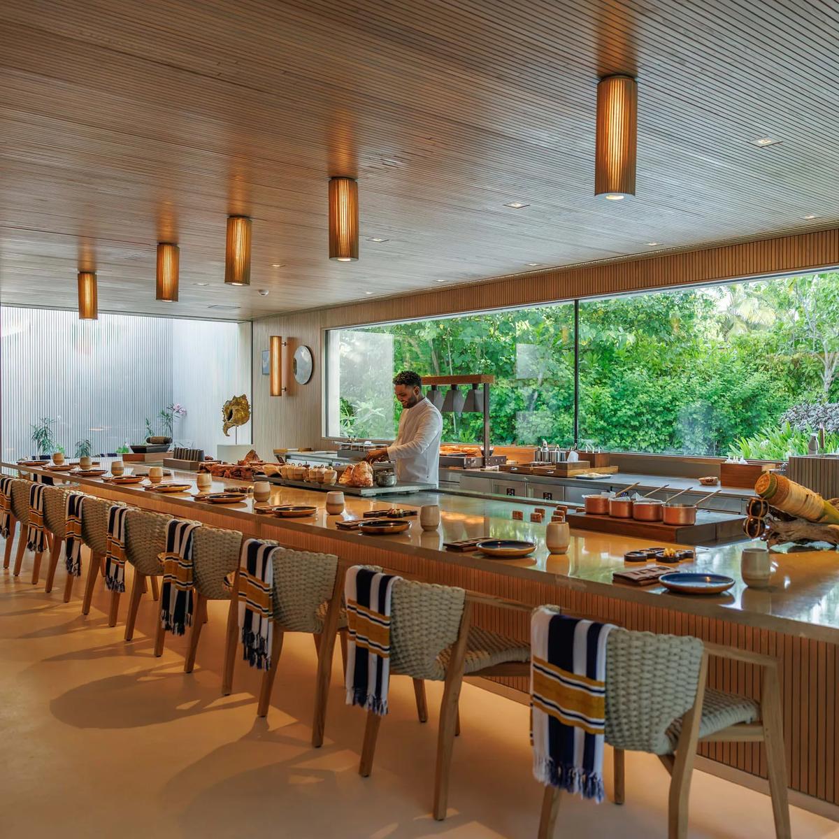 A chef preparing a dish at an open kitchen counter, floor-to-ceiling glass framing the garden behind him
