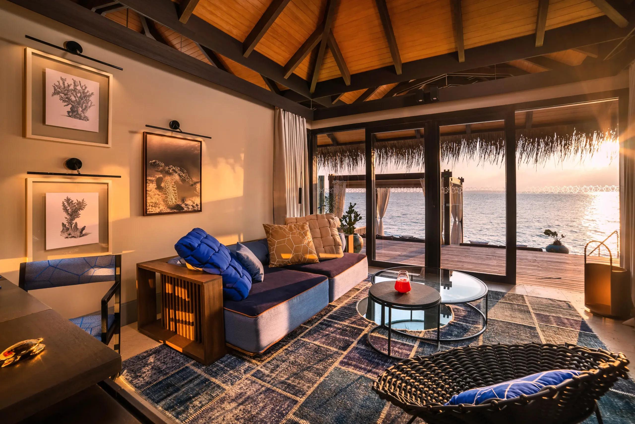 The living area of a Sunset Deluxe Water Pool Villa at dusk, glass doors open to an overwater deck and the sea beyond