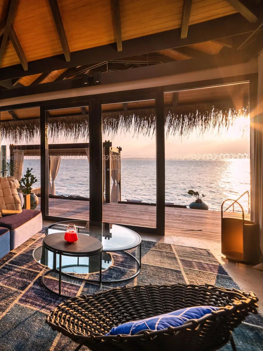 The living area of a Sunset Deluxe Water Pool Villa at dusk, glass doors open to an overwater deck and the sea beyond