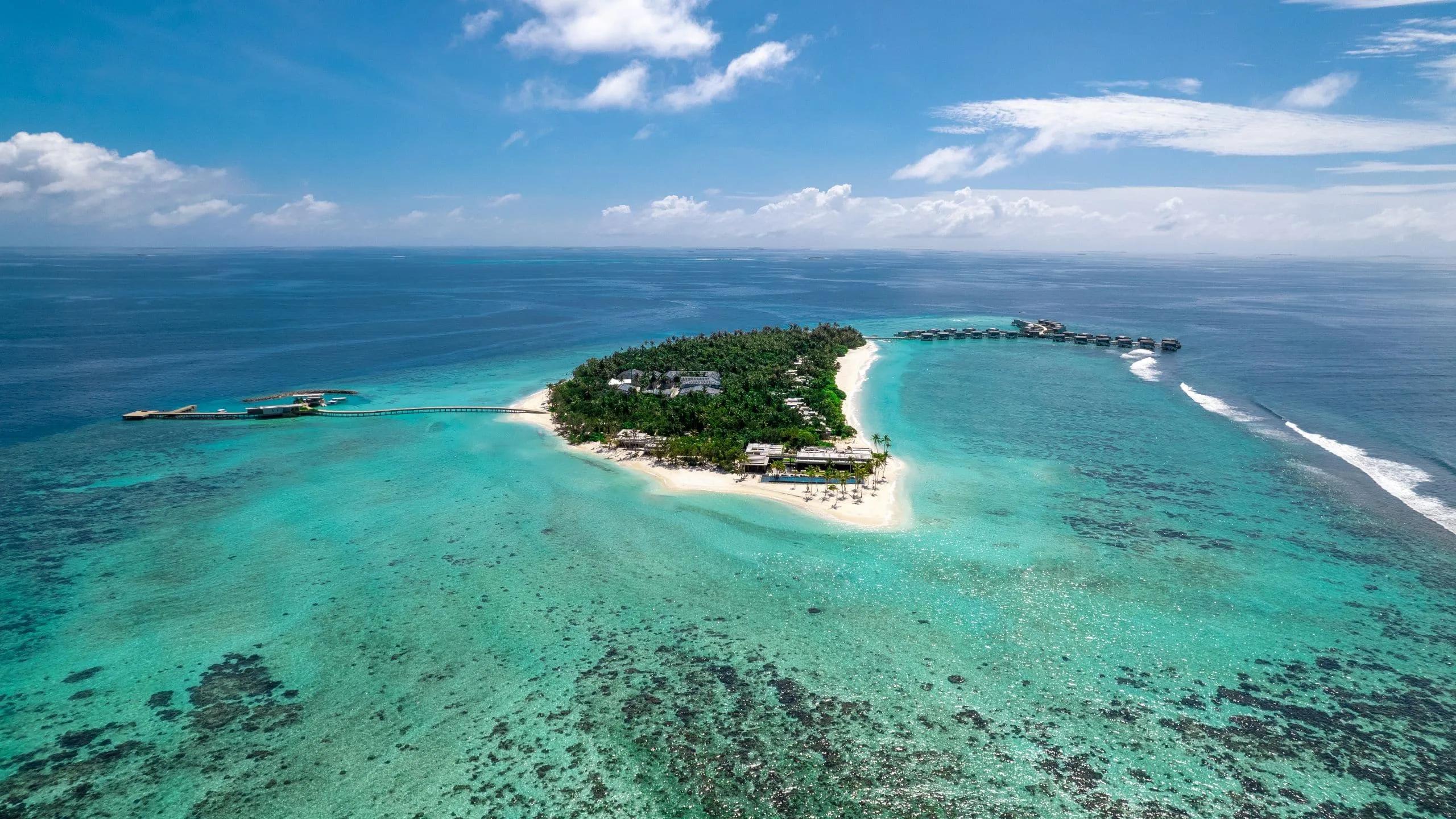 Alila Kothaifaru's island in Raa Atoll seen from the side, dense greenery and beach villas giving way to a row of overwater villas along the reef edge