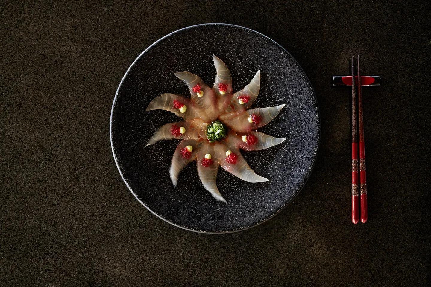 A sashimi preparation fanned into a star pattern on a black plate at Kanusan, garnished with roe and chives