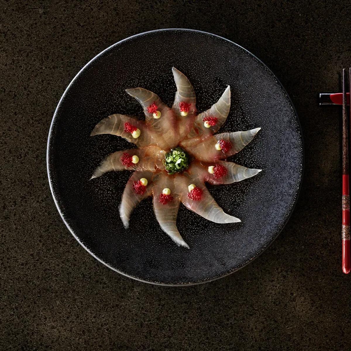 A sashimi preparation fanned into a star pattern on a black plate at Kanusan, garnished with roe and chives