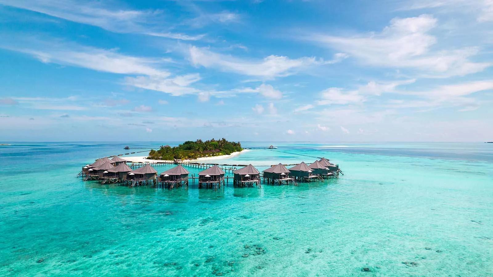 Aerial view of Komandoo's overwater villas curving off the reef's edge at sunrise, the island still in shadow behind them