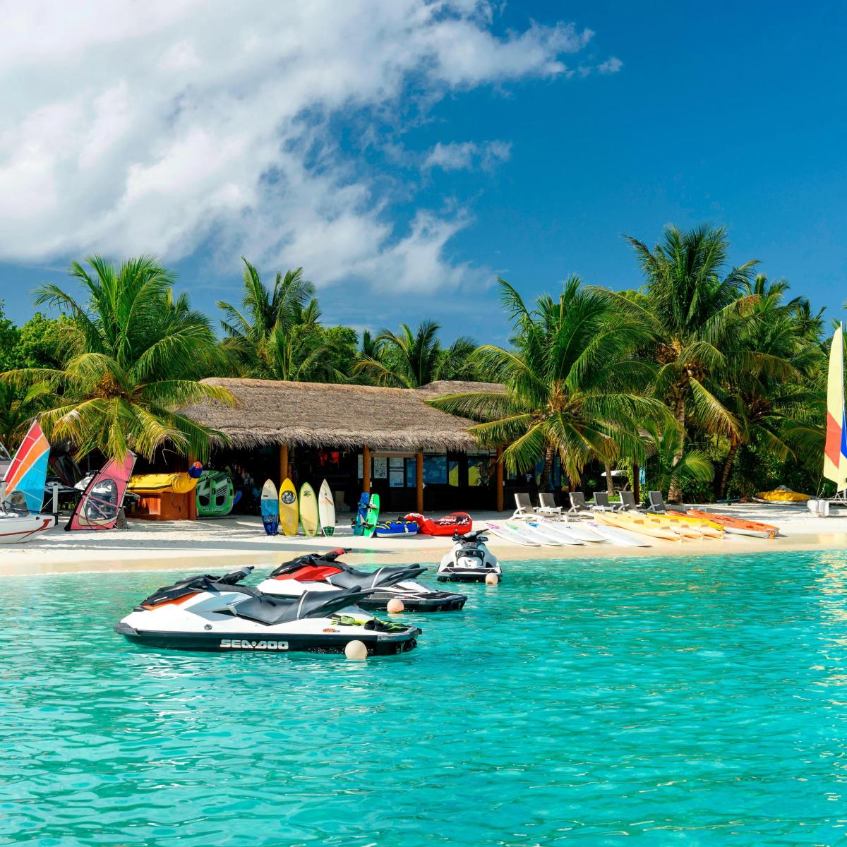 Kayaks, surfboards and jet skis lined up on the beach in front of the watersports hut, with sailboats moored just offshore
