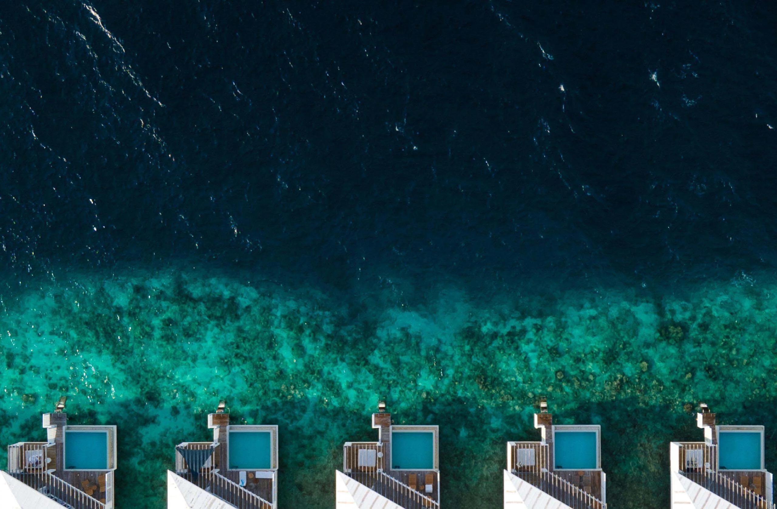 A row of overwater villas with plunge pools at Amilla, seen directly from above where the reef drops from turquoise to deep blue