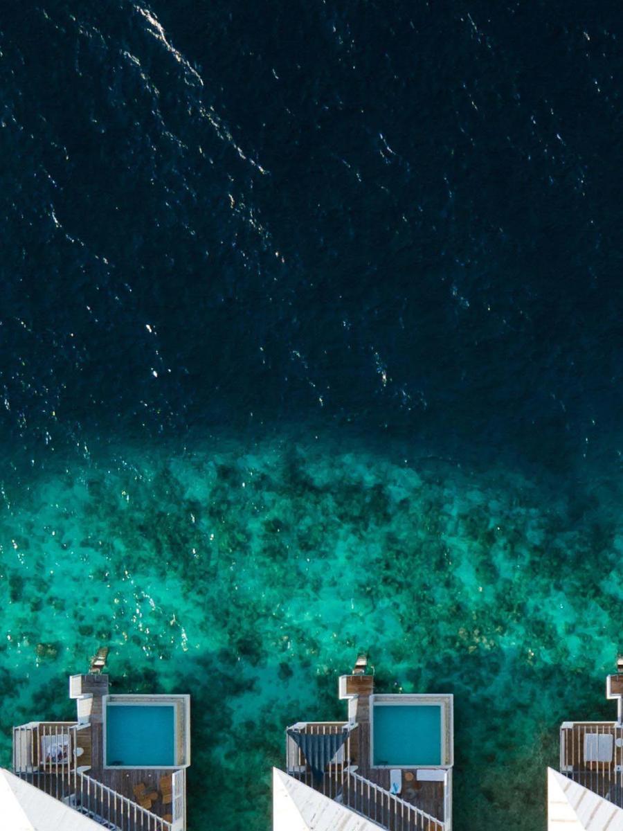 A row of overwater villas with plunge pools at Amilla, seen directly from above where the reef drops from turquoise to deep blue