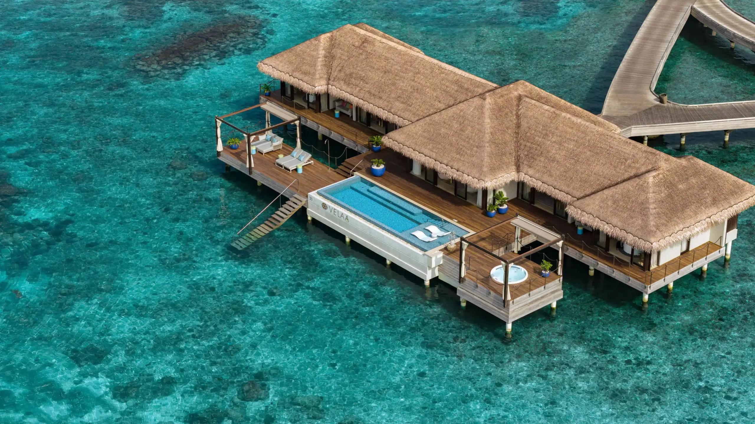 An aerial view of an overwater Ocean Pool House, its long infinity pool and thatched roofline set over the lagoon