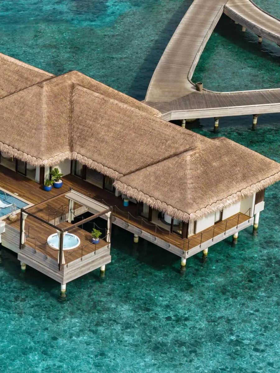 An aerial view of an overwater Ocean Pool House, its long infinity pool and thatched roofline set over the lagoon