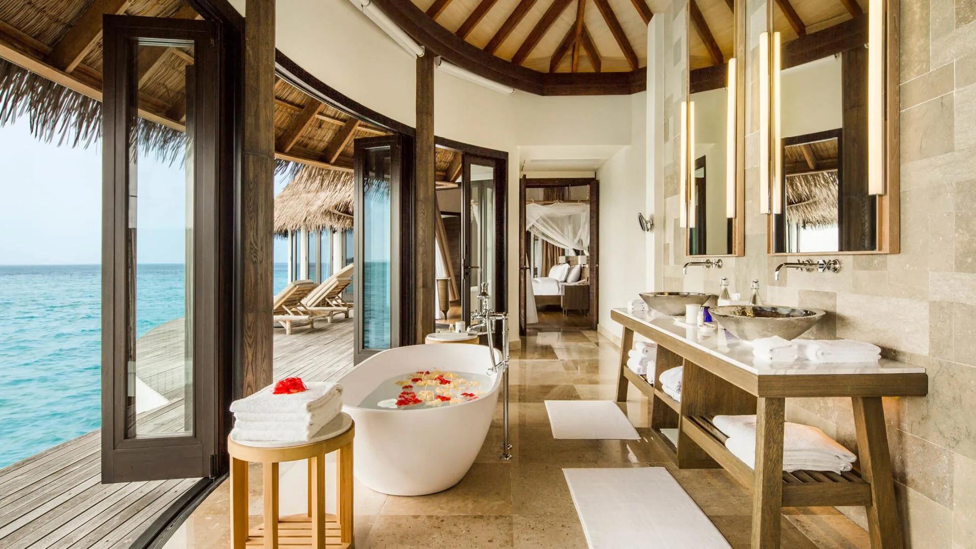 Freestanding bathtub filled with rose petals beside floor-to-ceiling windows opening onto an overwater deck and the ocean