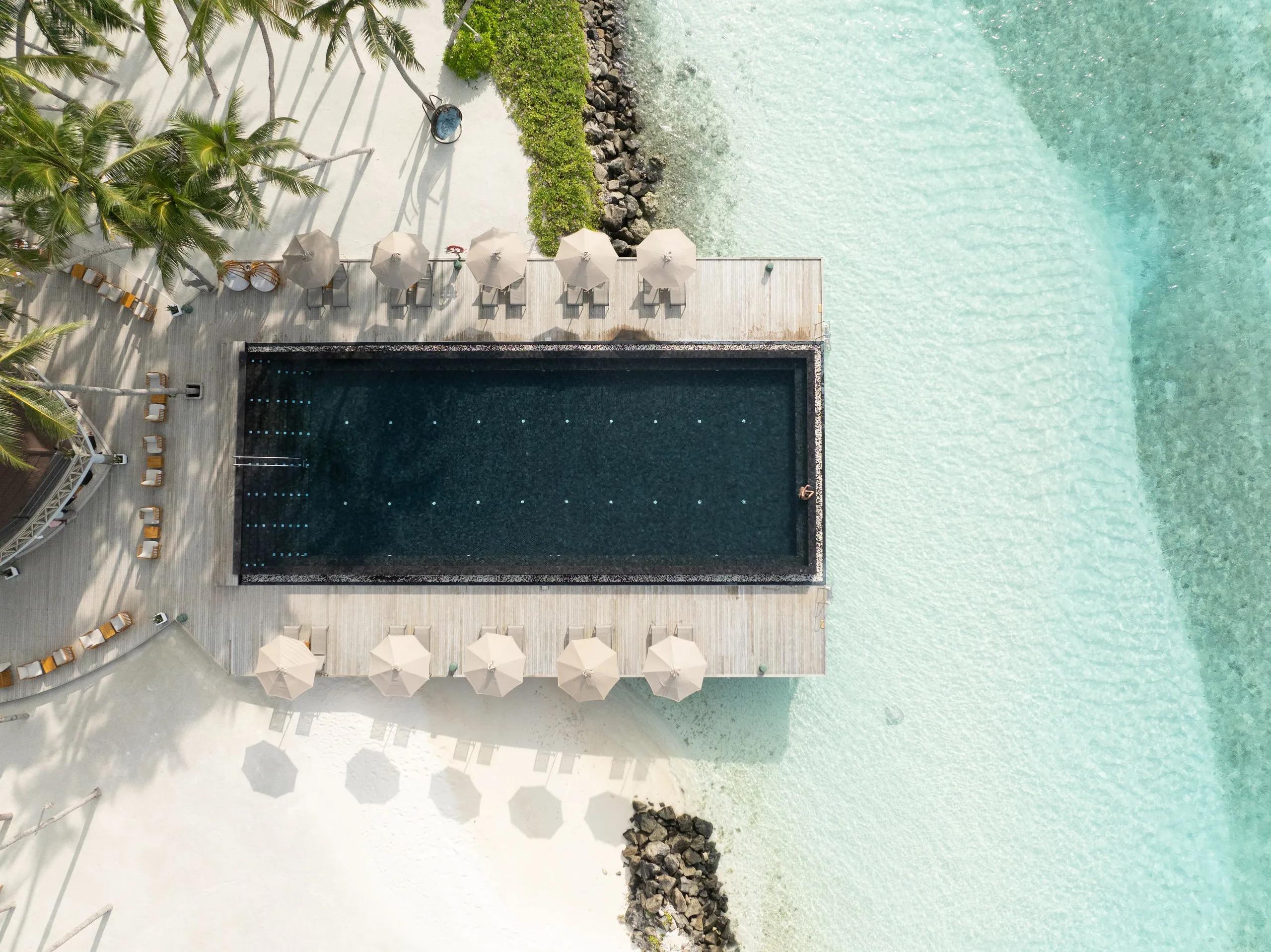 Aerial view straight down onto a dark overwater infinity pool surrounded by sun loungers and umbrellas, jutting out into the pale turquoise lagoon