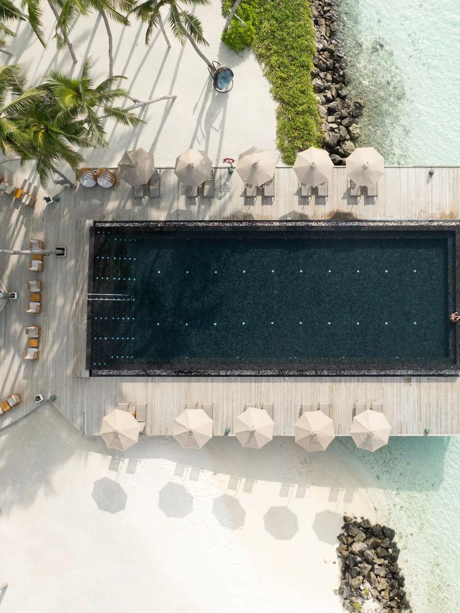 Aerial view straight down onto a dark overwater infinity pool surrounded by sun loungers and umbrellas, jutting out into the pale turquoise lagoon