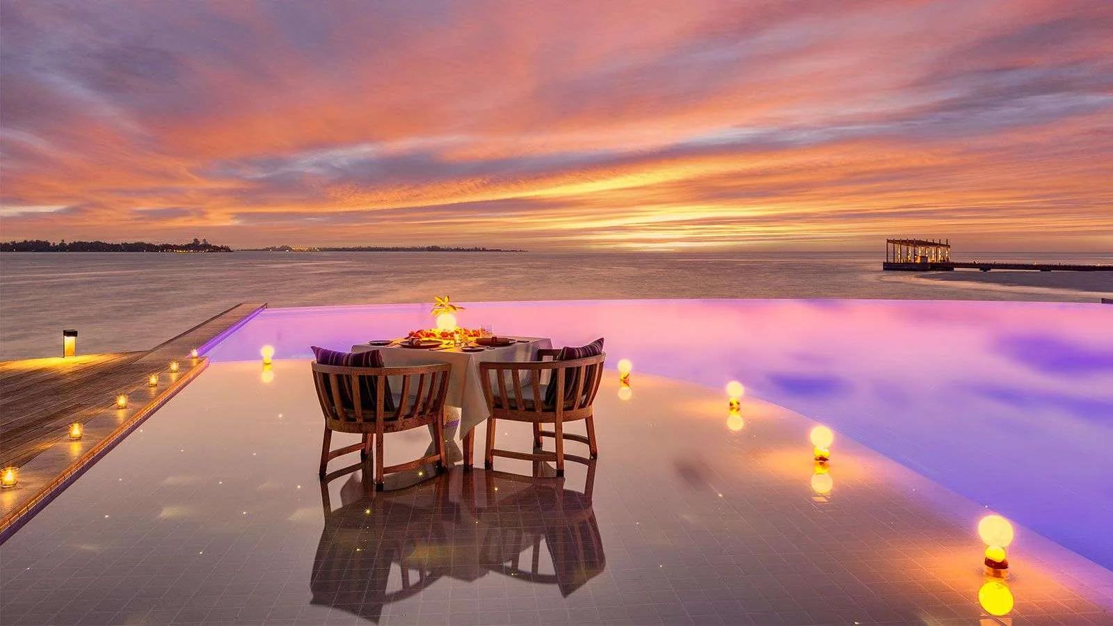 A candlelit table for two set at the edge of an infinity pool deck as the sky turns orange at dusk