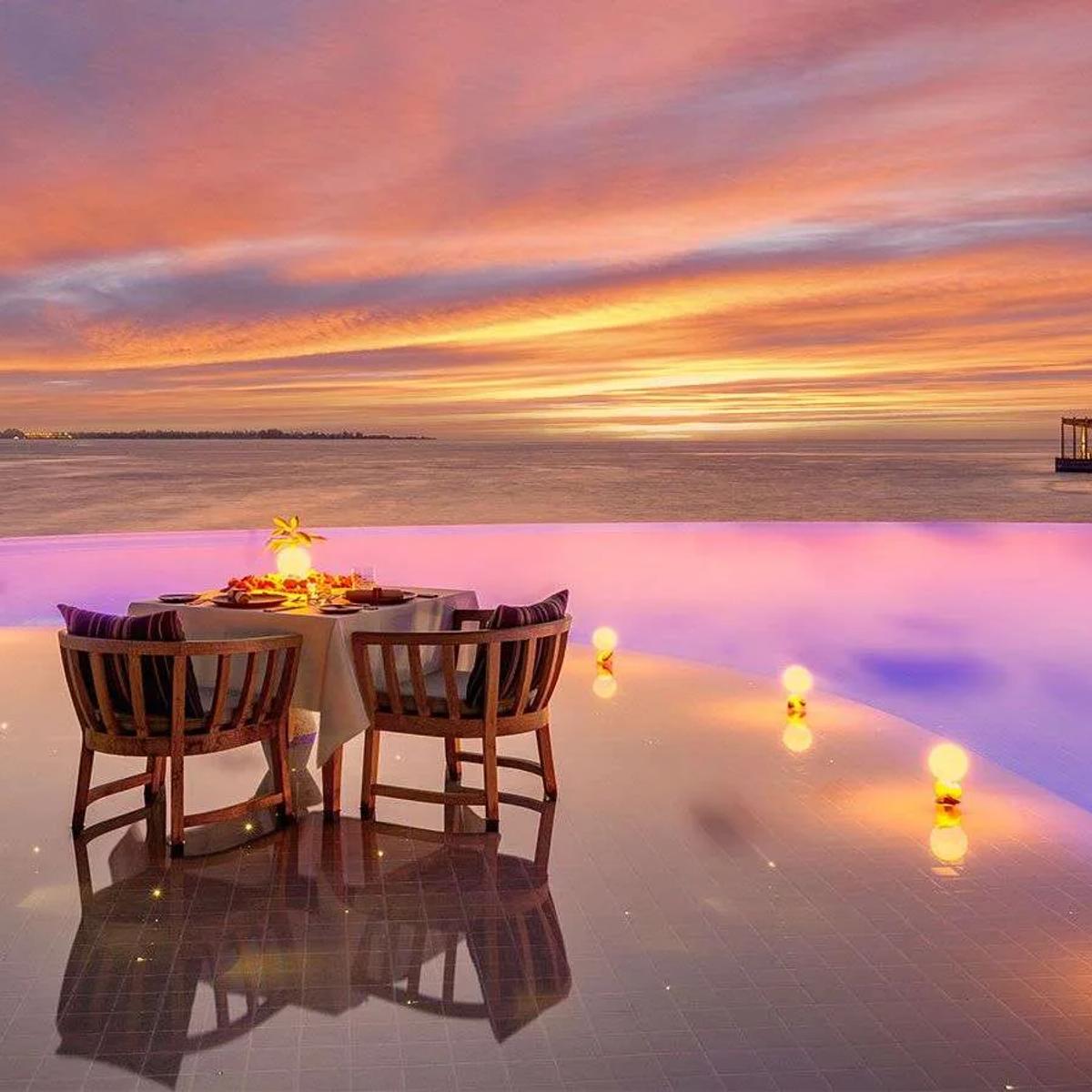 A candlelit table for two set at the edge of an infinity pool deck as the sky turns orange at dusk