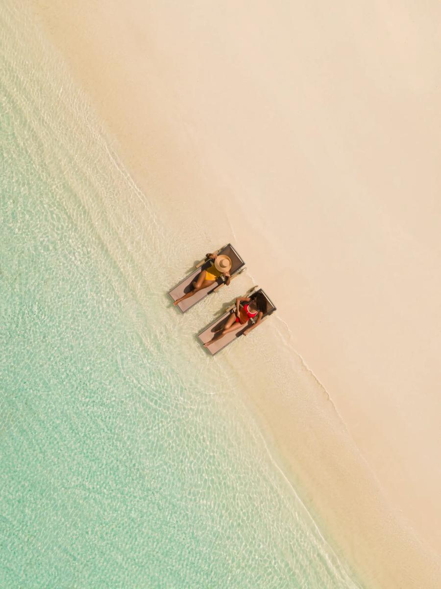 Two guests sunbathing on loungers under palm trees on Iru Fushi's beach, seen directly from above