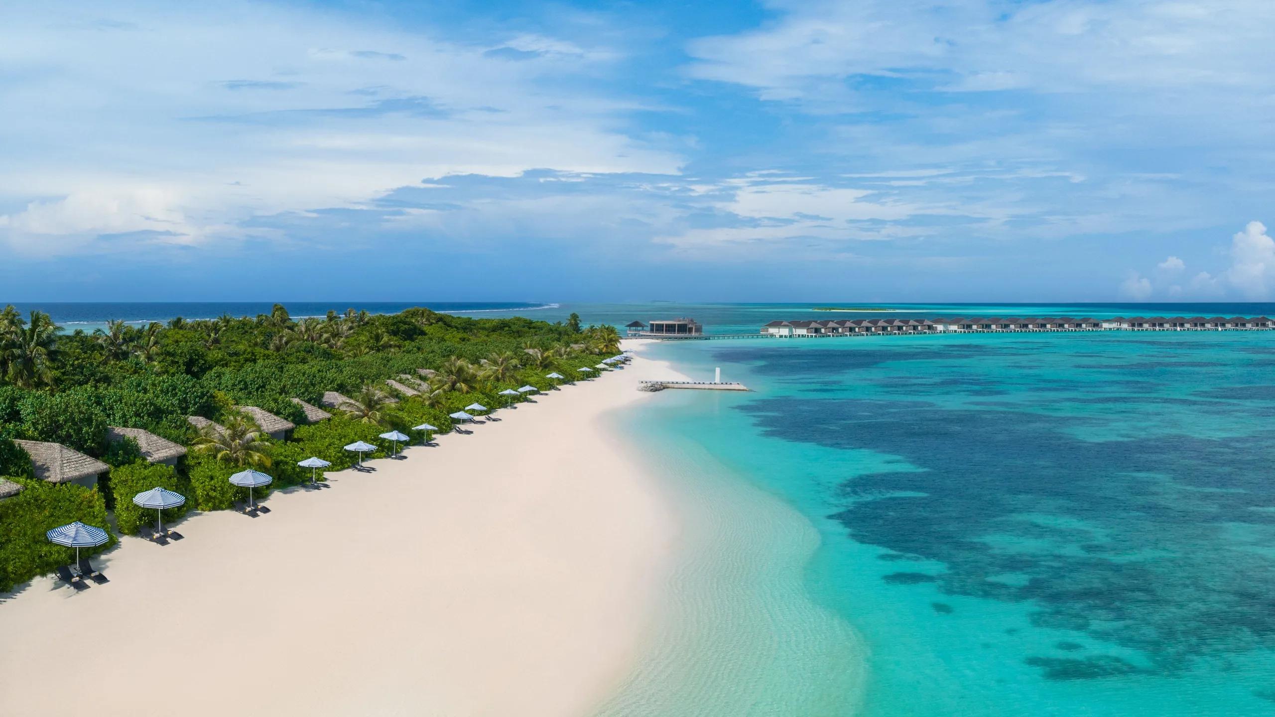 Aerial view of the beach, palm-lined shore and a row of overwater villas reaching into the lagoon at Le Méridien Maldives