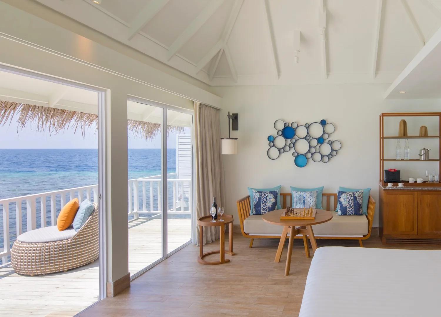 The living area of a Family Overwater Villa, daybed and chess table beside sliding doors onto the lagoon