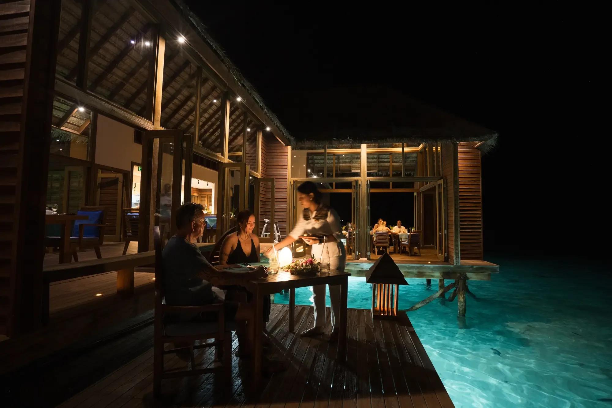 A couple dining at Mandhoo Restaurant's overwater deck at night, lit by lantern light at Conrad Maldives Rangali
