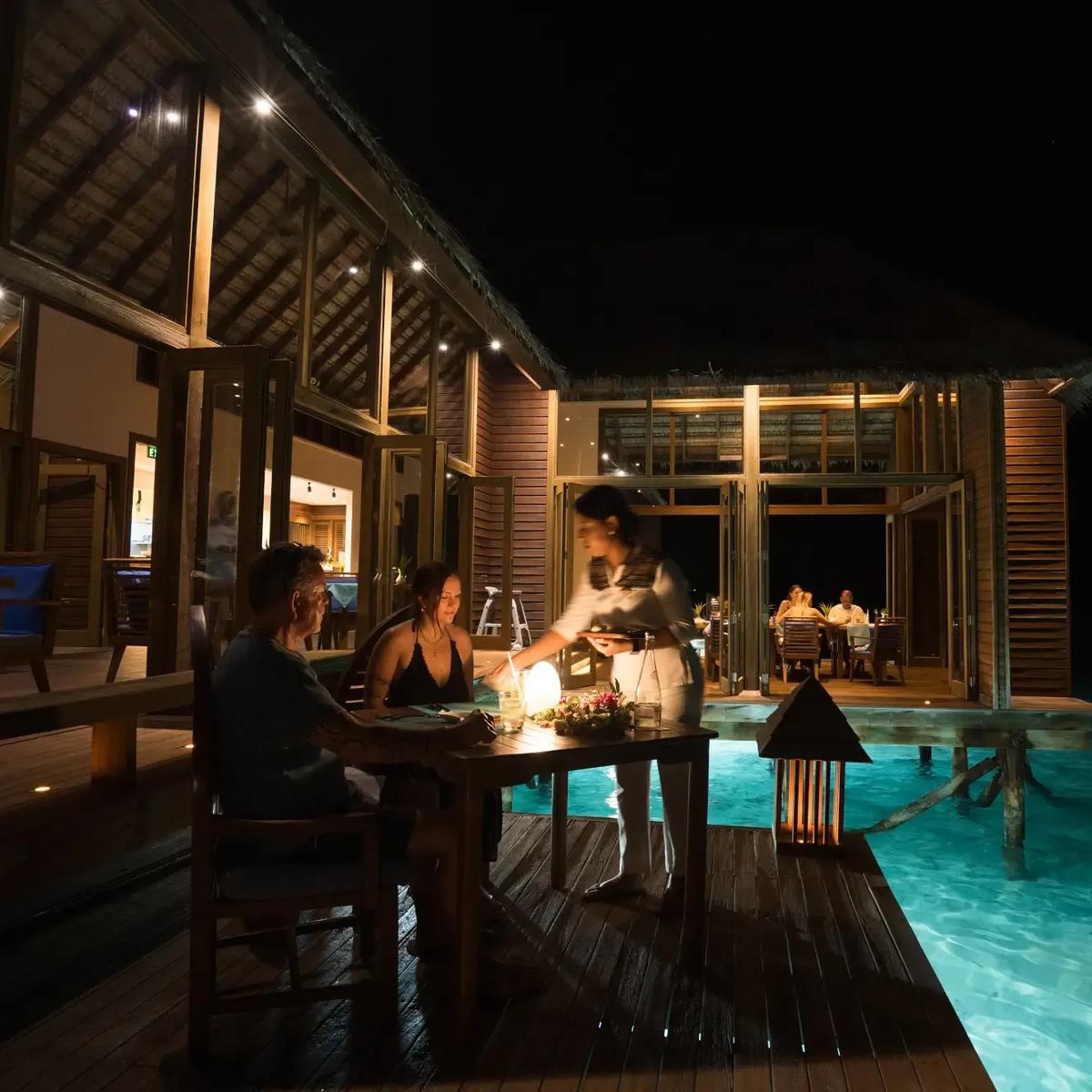 A couple dining at Mandhoo Restaurant's overwater deck at night, lit by lantern light at Conrad Maldives Rangali
